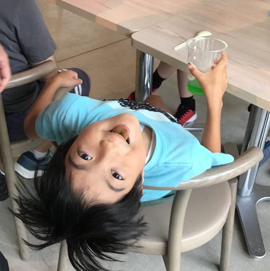 Child lying on a chair with his head hanging off the edge, smiling at the camera as friends sit at a table nearby with drinks.