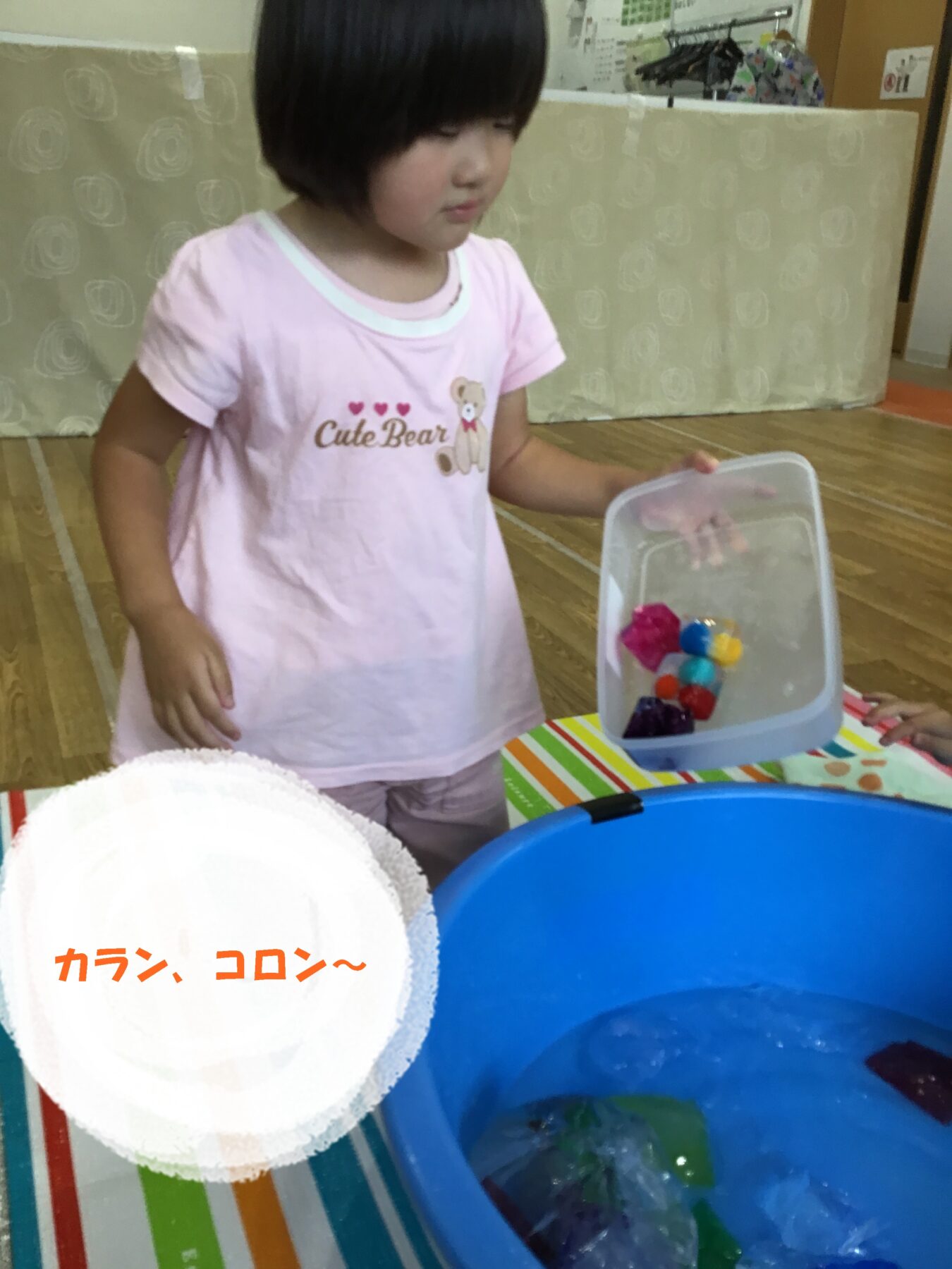 Young girl in a pink shirt with a bear graphic and 'Cute Bear' text stands by a blue tub of water, holding a clear plastic box of colorful toys/objects.