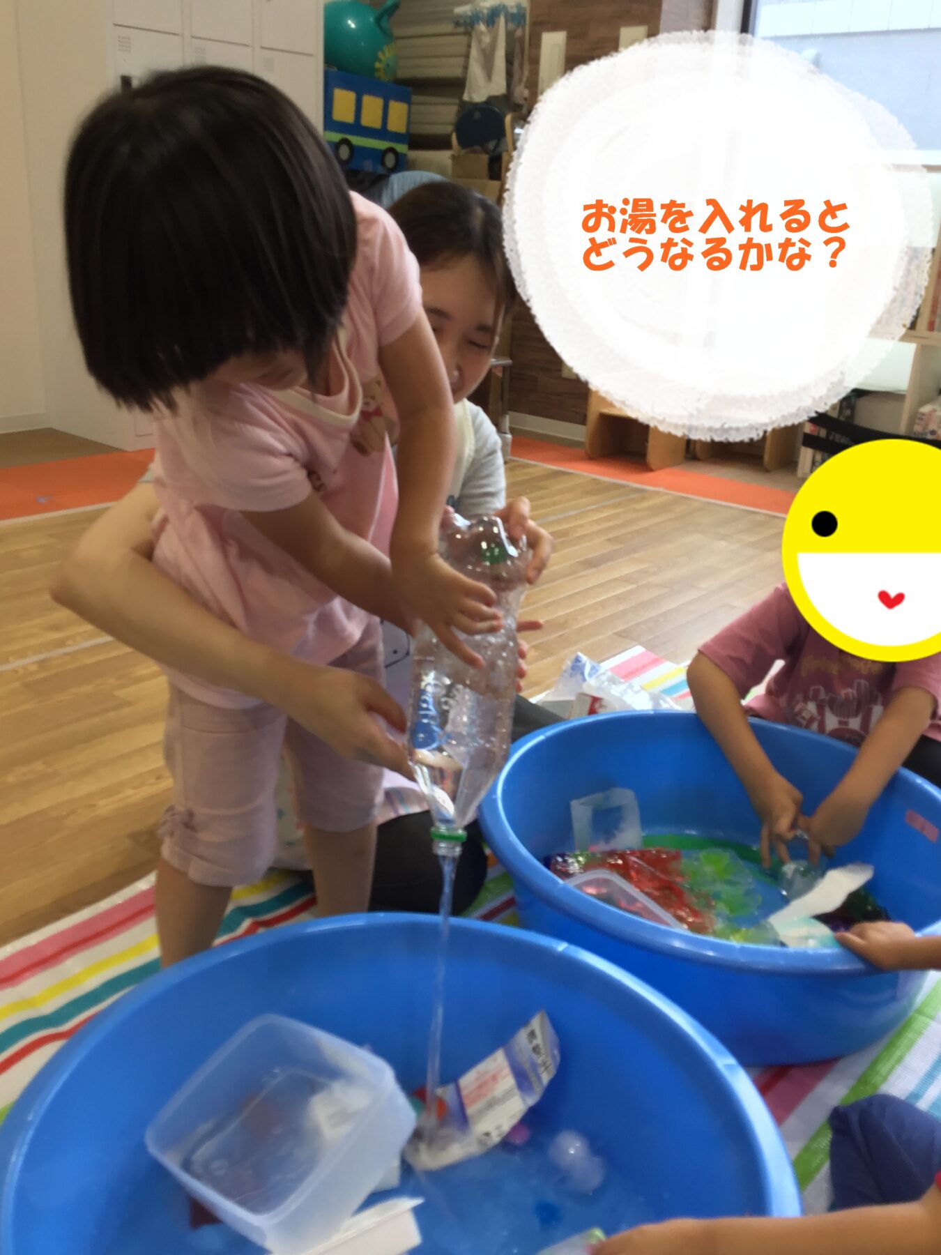 Two children pour water into a blue tub during a classroom activity, with bags and supplies nearby on a colorful mat inside a room.