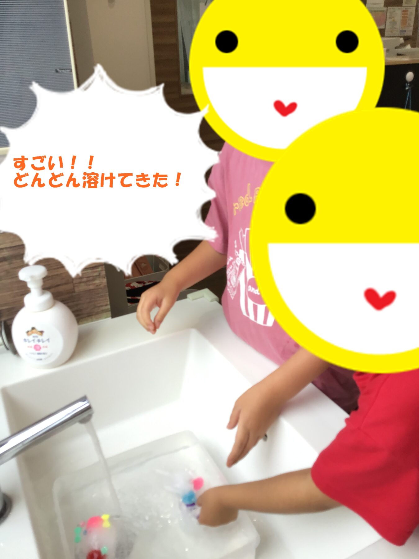 Two kids at a bathroom sink playing with colorful beads in soapy water, their faces covered by large yellow emoji stickers.