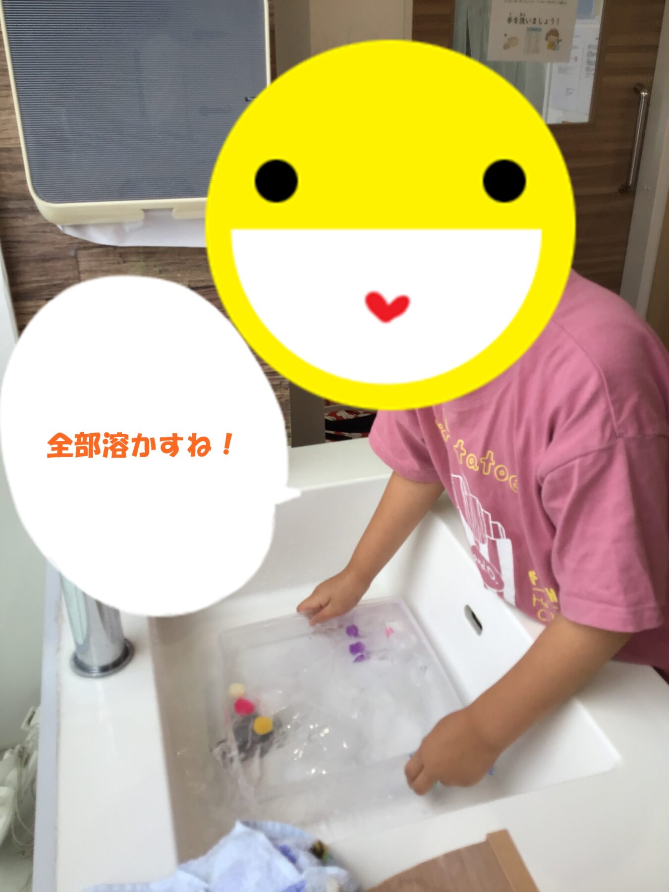 Child in a pink shirt washes hands at a sink; a large yellow smiley overlay covers most of the face area, with a white speech bubble on the left containing orange Japanese text.