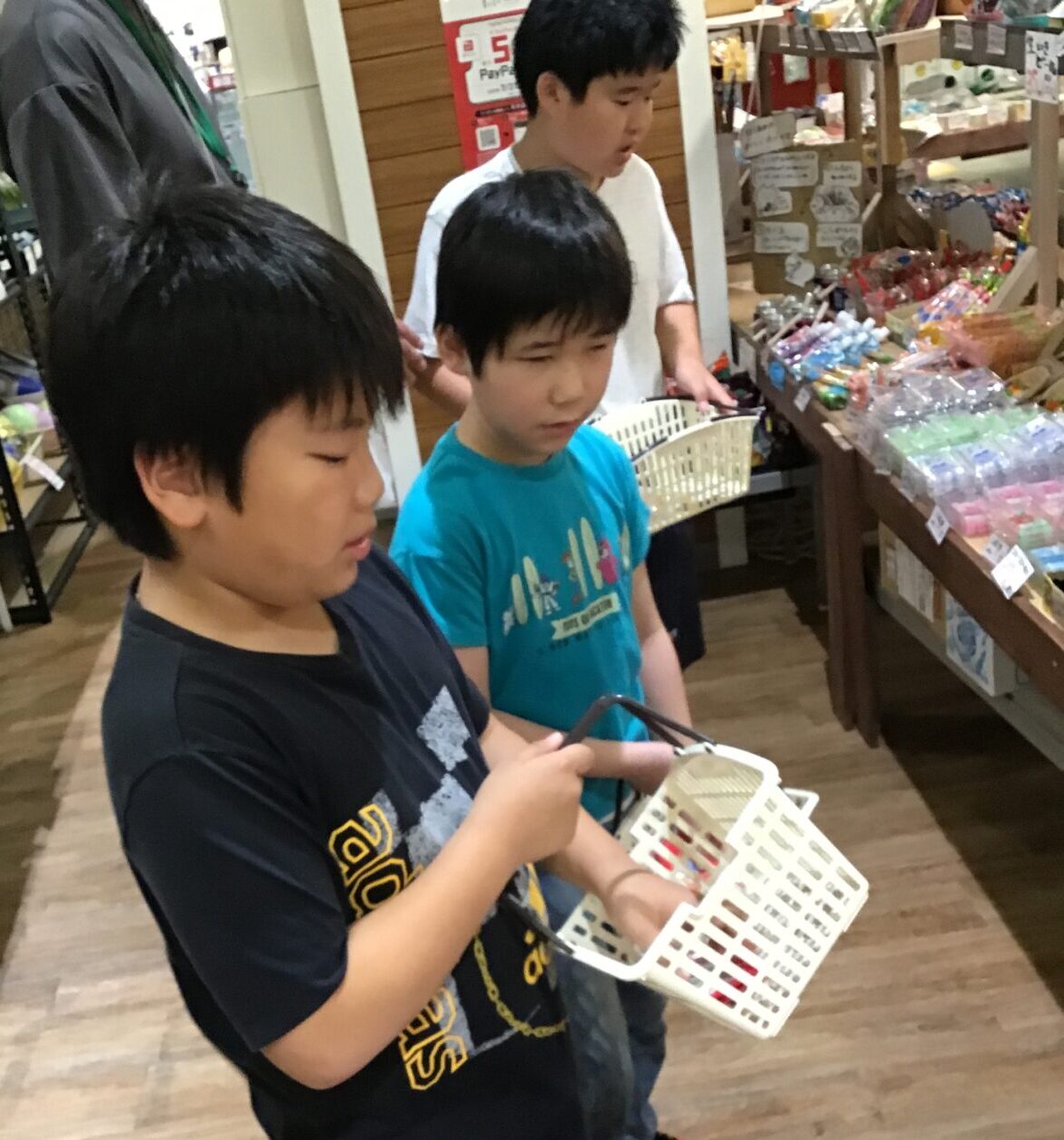 Three boys in a store, each with a small basket, choosing items from a candy display.