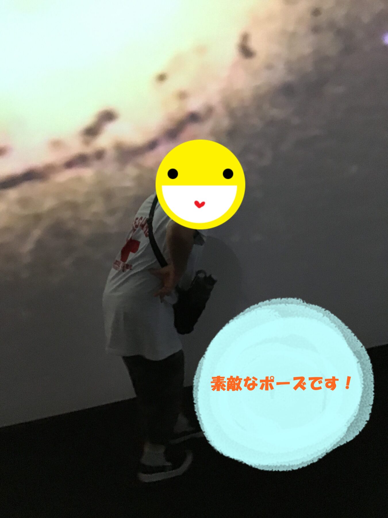 Person posing on a dark stage with their face covered by a large yellow emoji sticker; a glowing blue circle with orange Japanese text sits in front