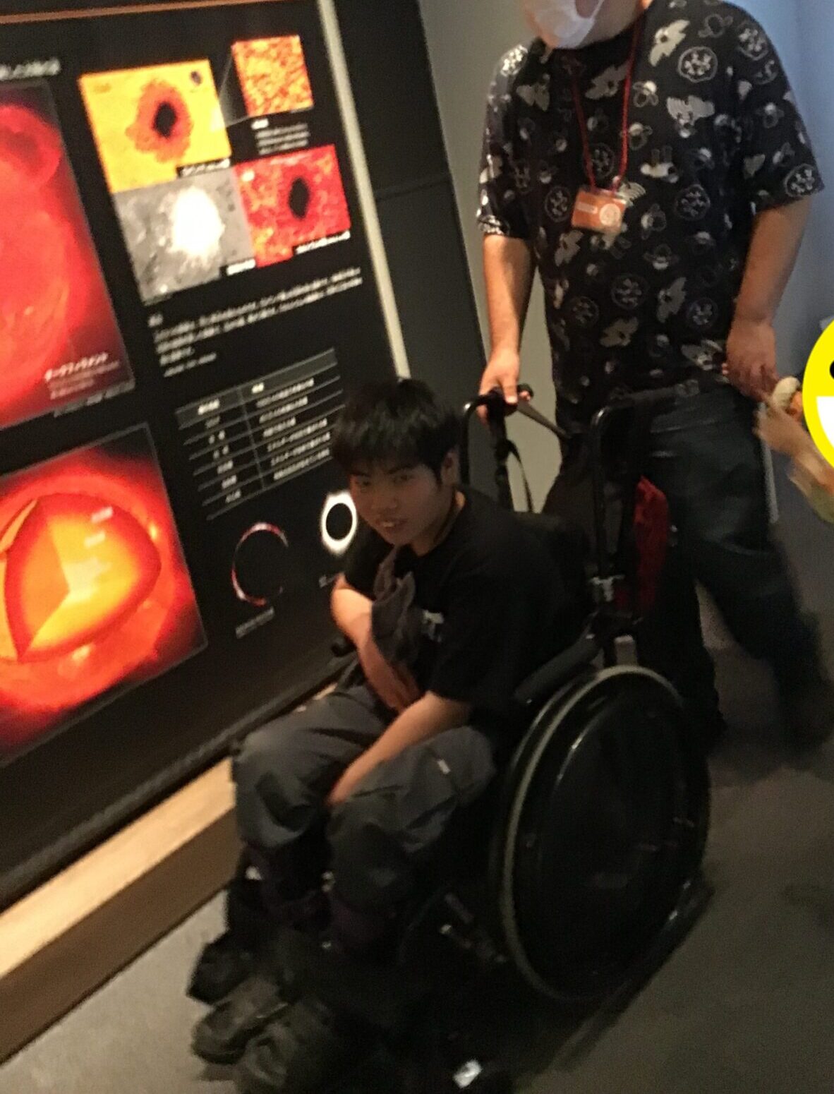 Person in a patterned shirt and mask assisting a young person in a wheelchair beside a museum exhibit with orange artwork panels.