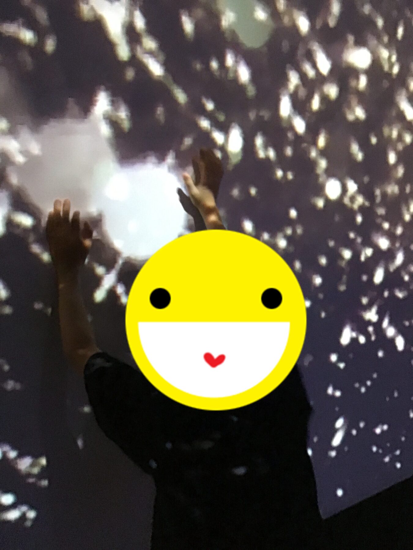 Person raises both hands in the air against a background of scattered bright lights. A large yellow smiley sticker covers the person’s face.
