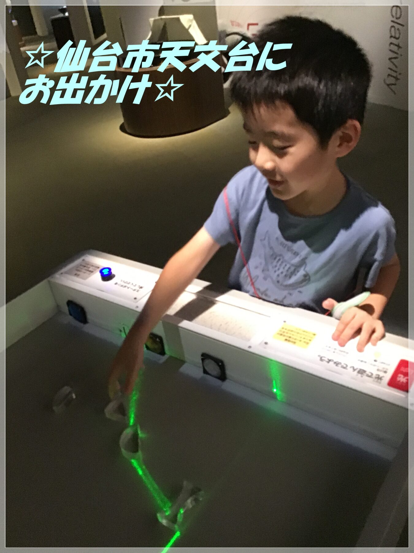 Young boy touching a control panel of an arcade exhibit; a green laser line glows on the floor and blue Japanese text overlays the scene.