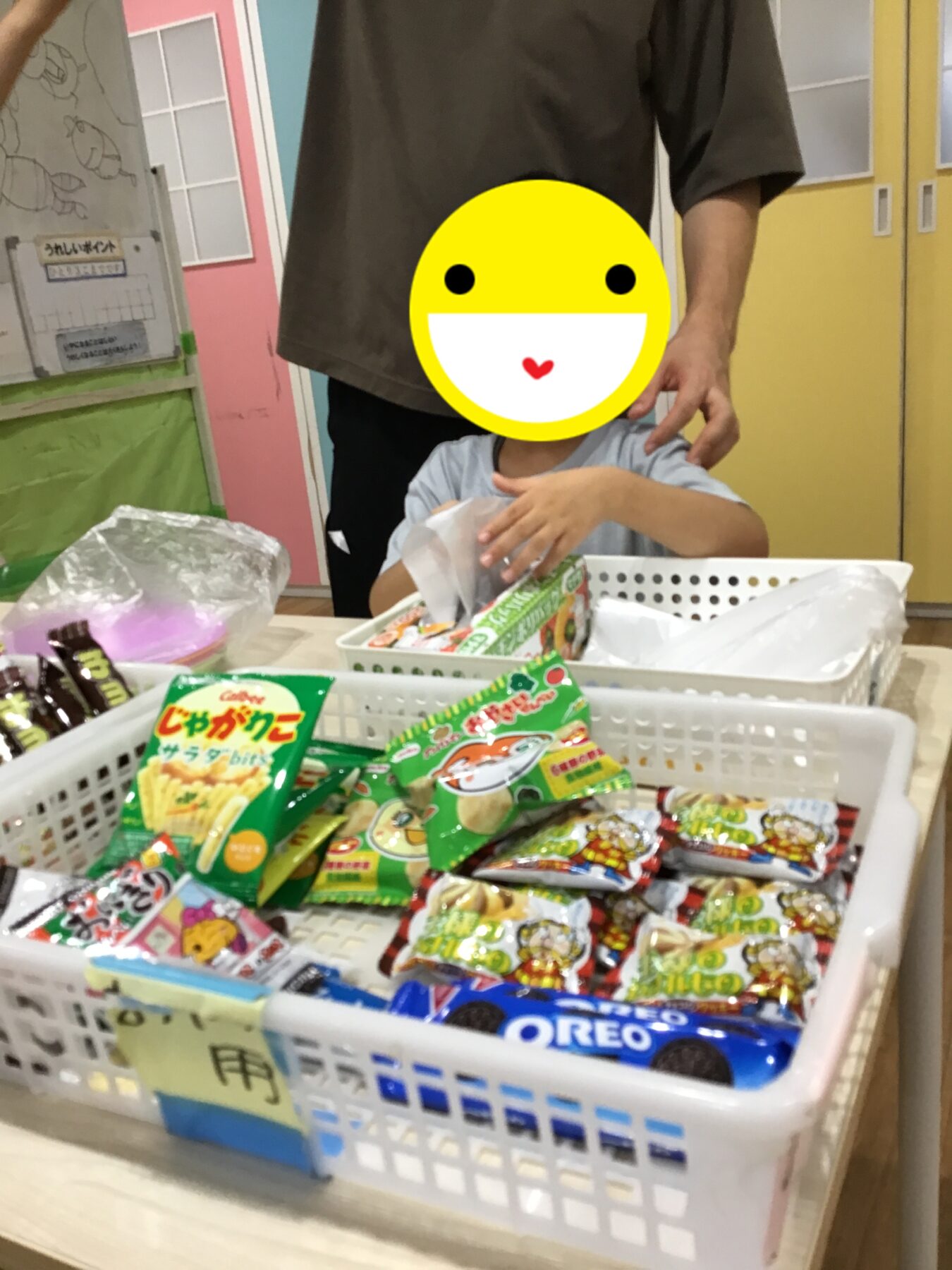 Child standing at a table of colorful snack packets in baskets, with an adult's hand resting on their shoulder.