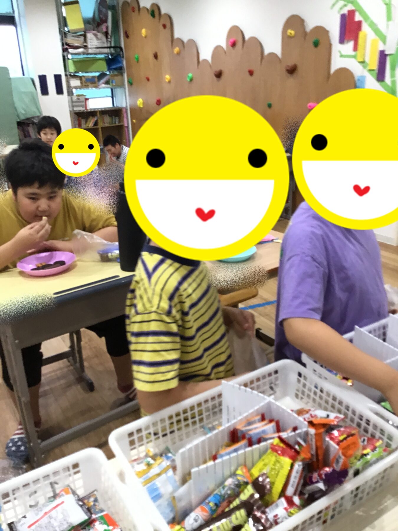 Children at a classroom snack activity, sorting and picking candy from white baskets on a table, with a colorful wall in the background and smiley-face stickers covering faces for privacy.