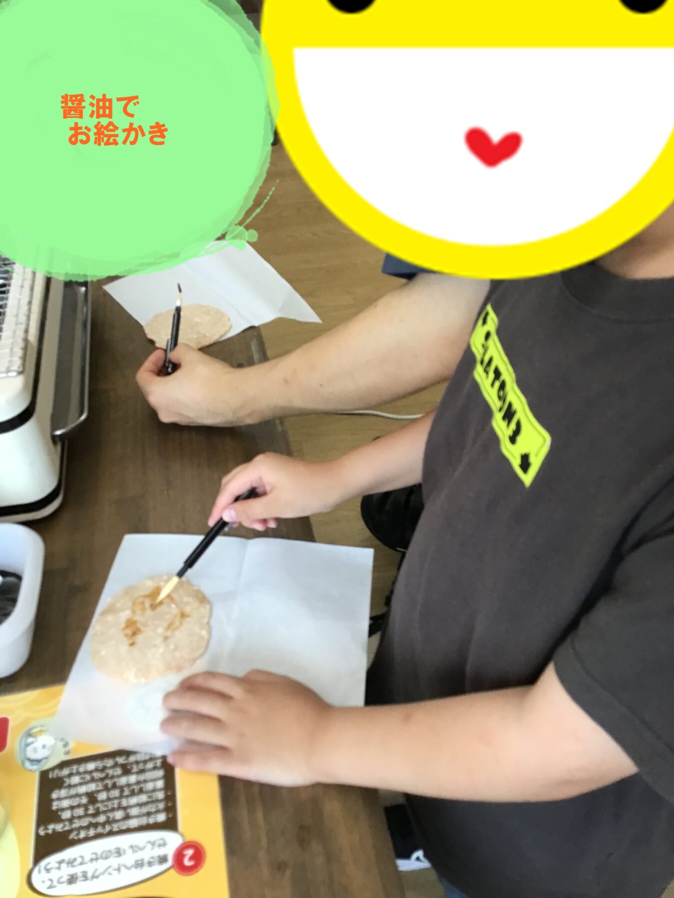 Person brushing sauce onto a round rice cracker on parchment at a table; emoji overlay covers the face.