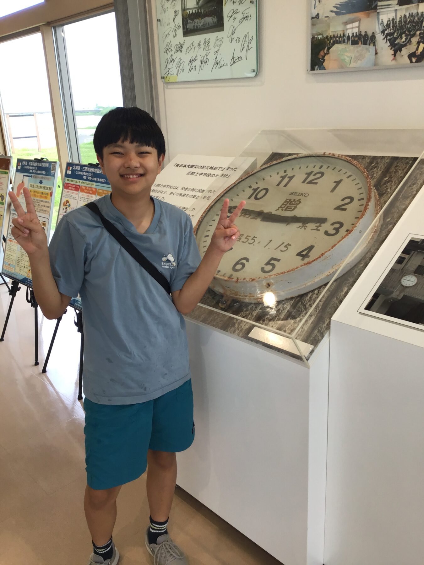 Smiling boy in a light blue shirt and turquoise shorts poses with peace signs next to a large tilted clock display in a museum-like space.