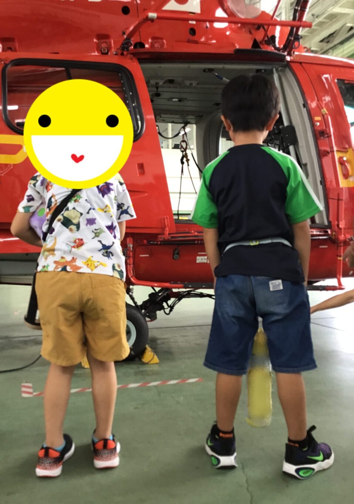 Three kids stand in a hangar beside a red helicopter with an open side door; one child’s face is covered by a large yellow smiley sticker.