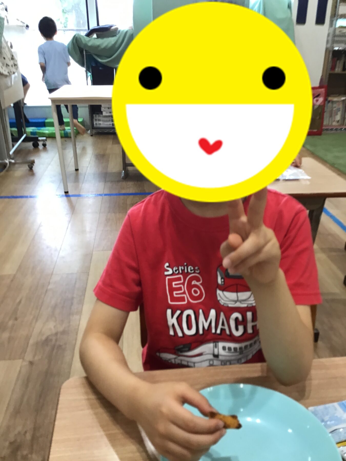 Child in a red shirt sits at a table, making a peace sign; a large yellow smiley overlay covers their face, with a blue plate in the foreground.