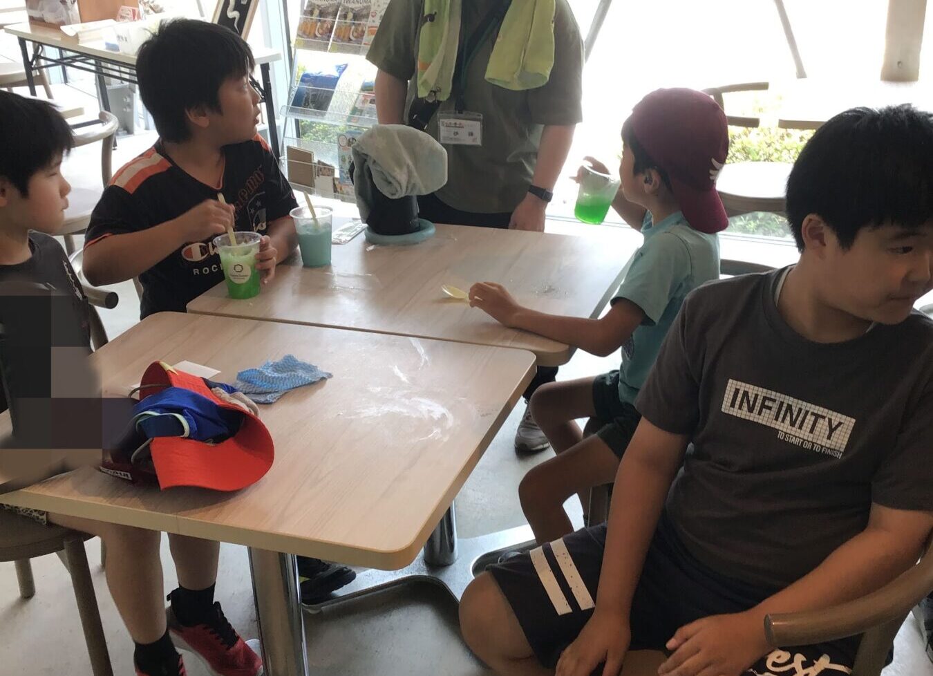 Instructor in a bucket hat talks to four children at a cafeteria-style table with colorful drinks; bright windowed room in the background.