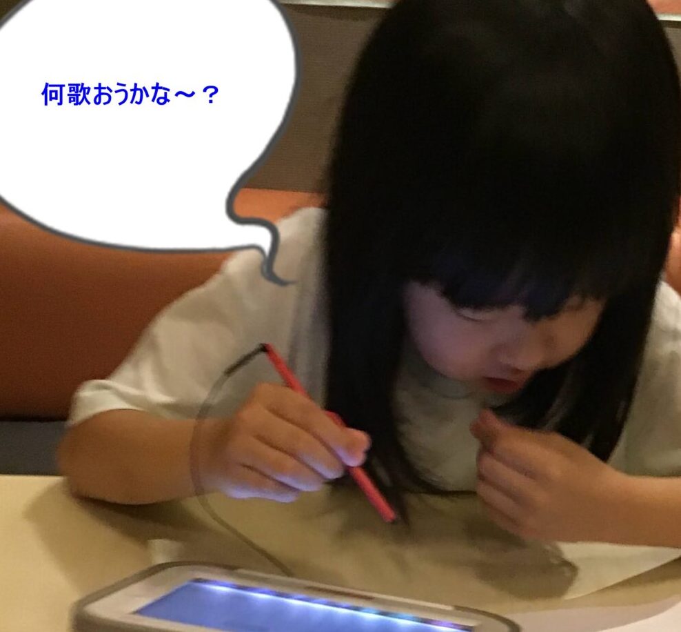 Young girl writes with a red pencil on a tablet, with papers containing Japanese text on the table in front of her.
