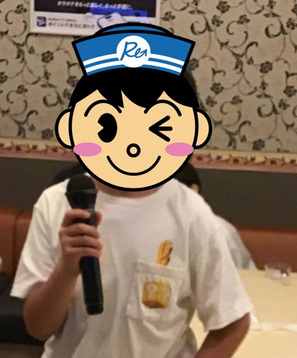 Child in a white T-shirt holding a microphone, standing in a restaurant with patterned wallpaper in the background.