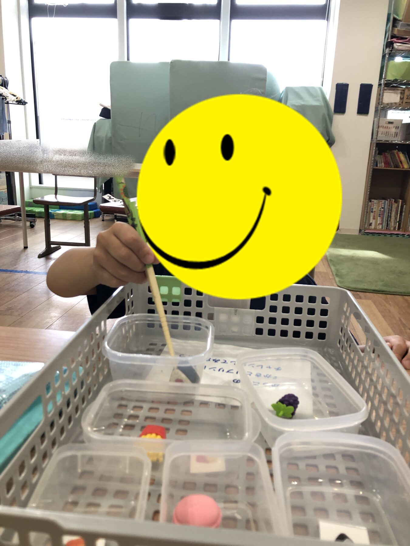 Child with a yellow smiley mask using a chopstick to explore small containers in a basket-filled craft area.