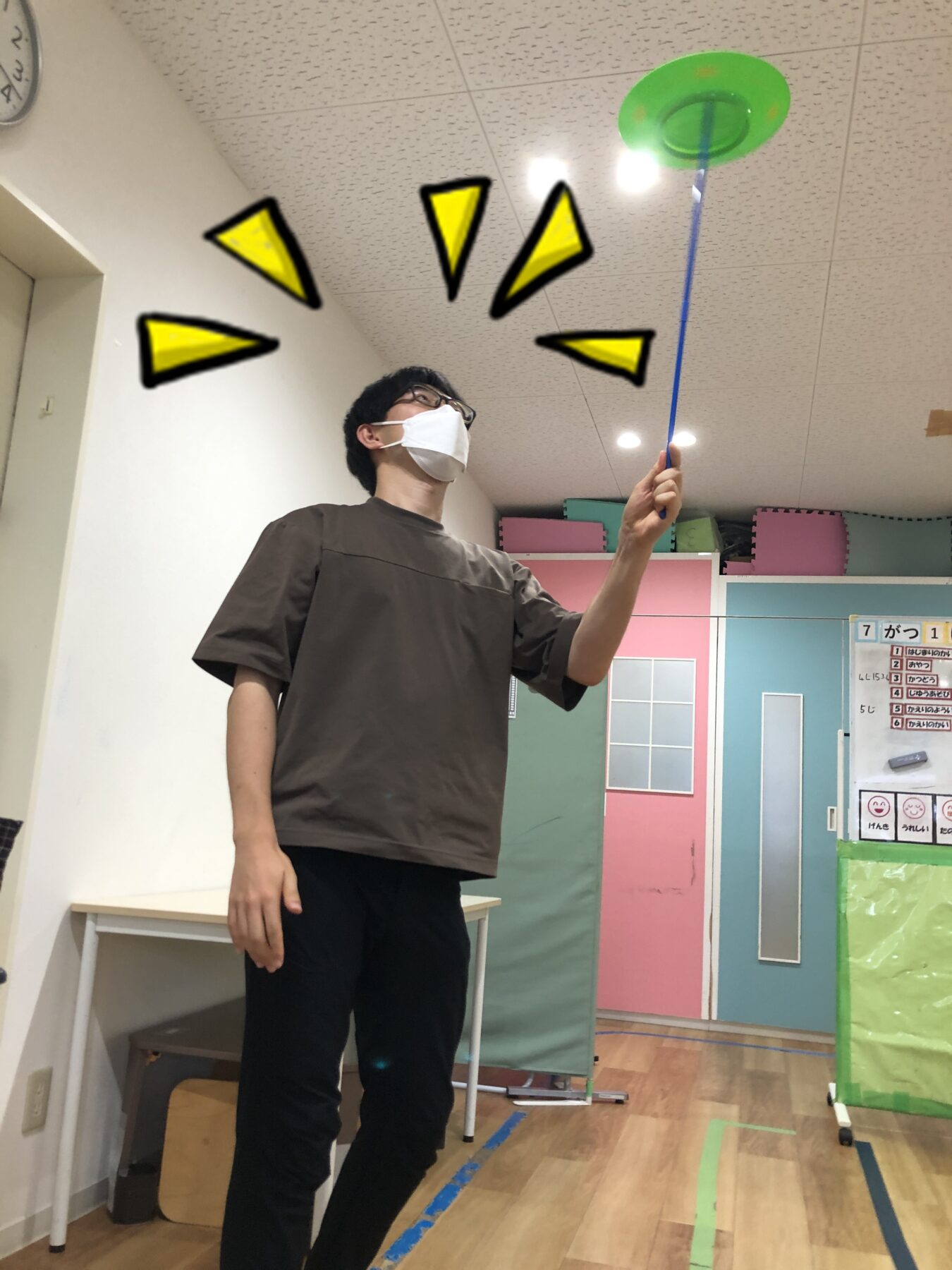 Man wearing a white face mask lifts a blue rod with a green frisbee at the end, indoors in a colorful room.