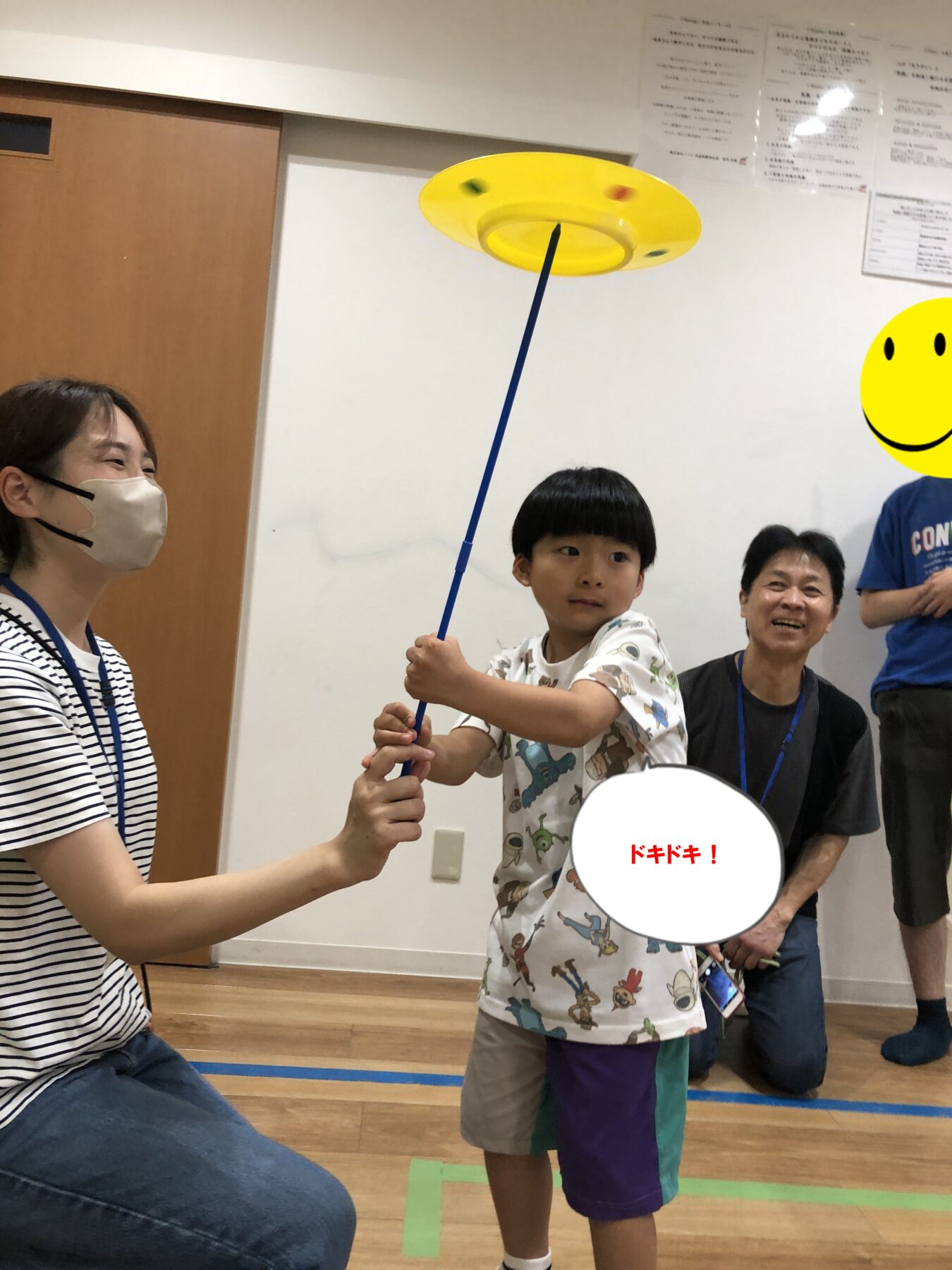 Boy holding a blue stick with a yellow tetherball spinning on top, guided by a masked woman; smiling adults watch in a room.