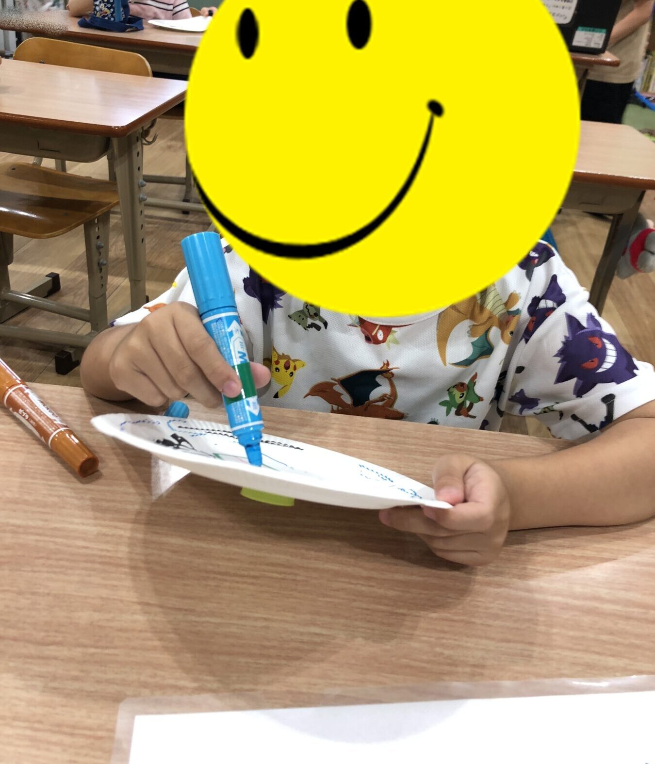 Child at a classroom desk drawing with a blue marker on a paper plate; large yellow smiley-face stickers cover faces, with classmates in the background.