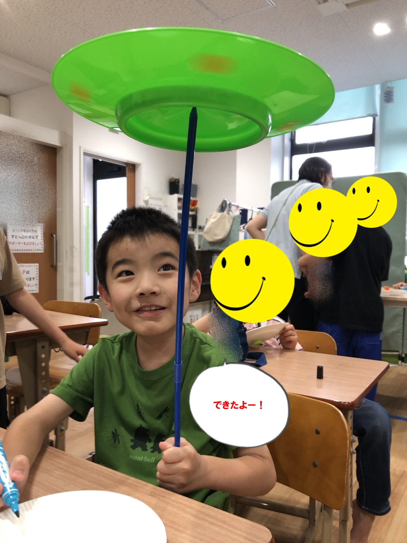 Boy in a green shirt holding a blue stick with a large green frisbee overhead in a classroom; smiling kids and smiley decorations in the background.