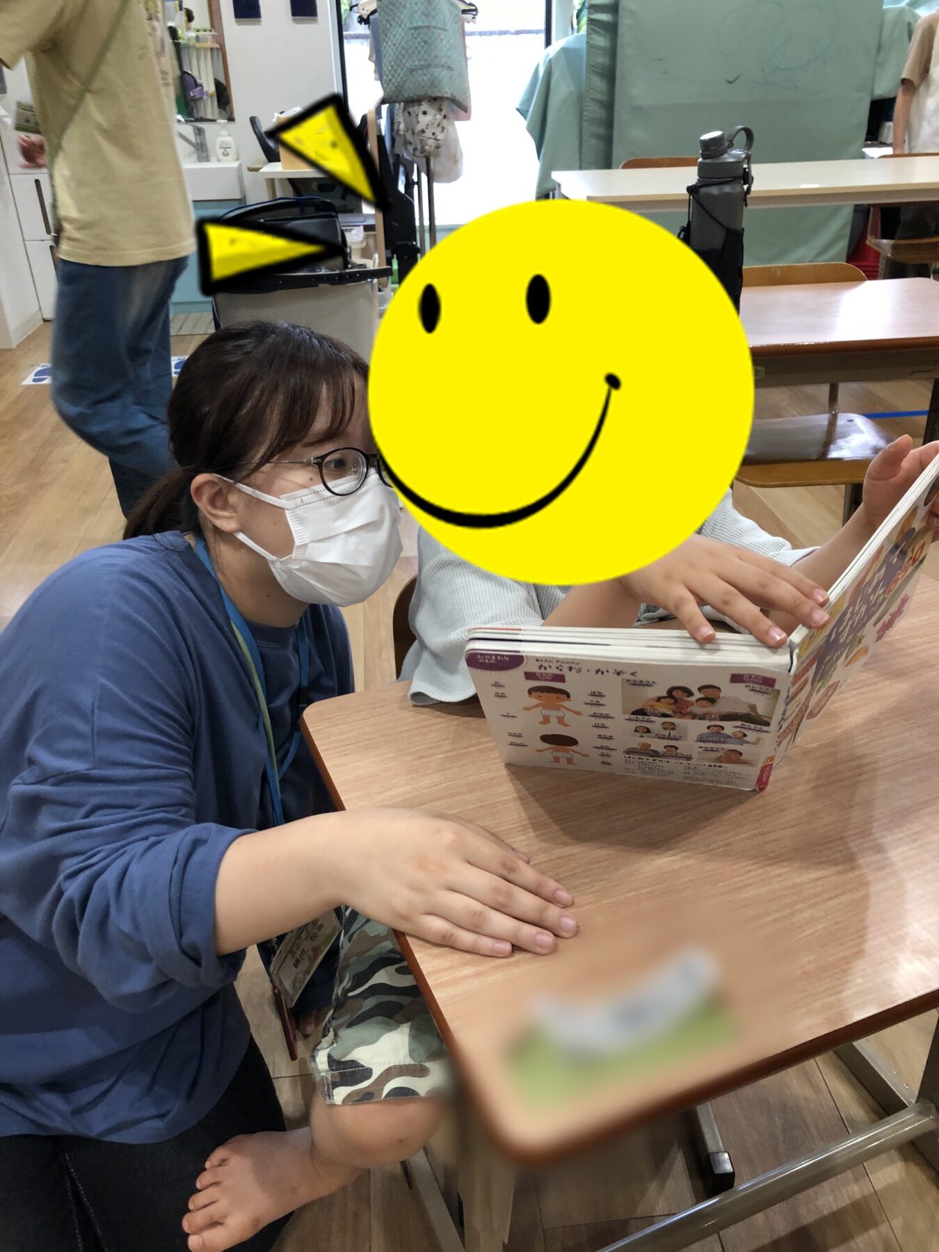 Caregiver in blue scrubs wearing a mask sits beside a child at a table, reading a colorful picture book together.