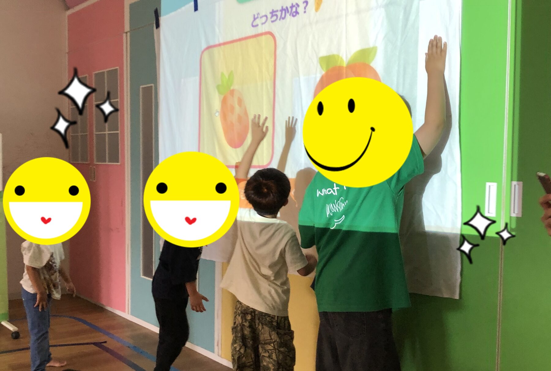 Children raise hands at a large colorful wall panel with a fruit illustration, in a classroom activity about choice or guessing.
