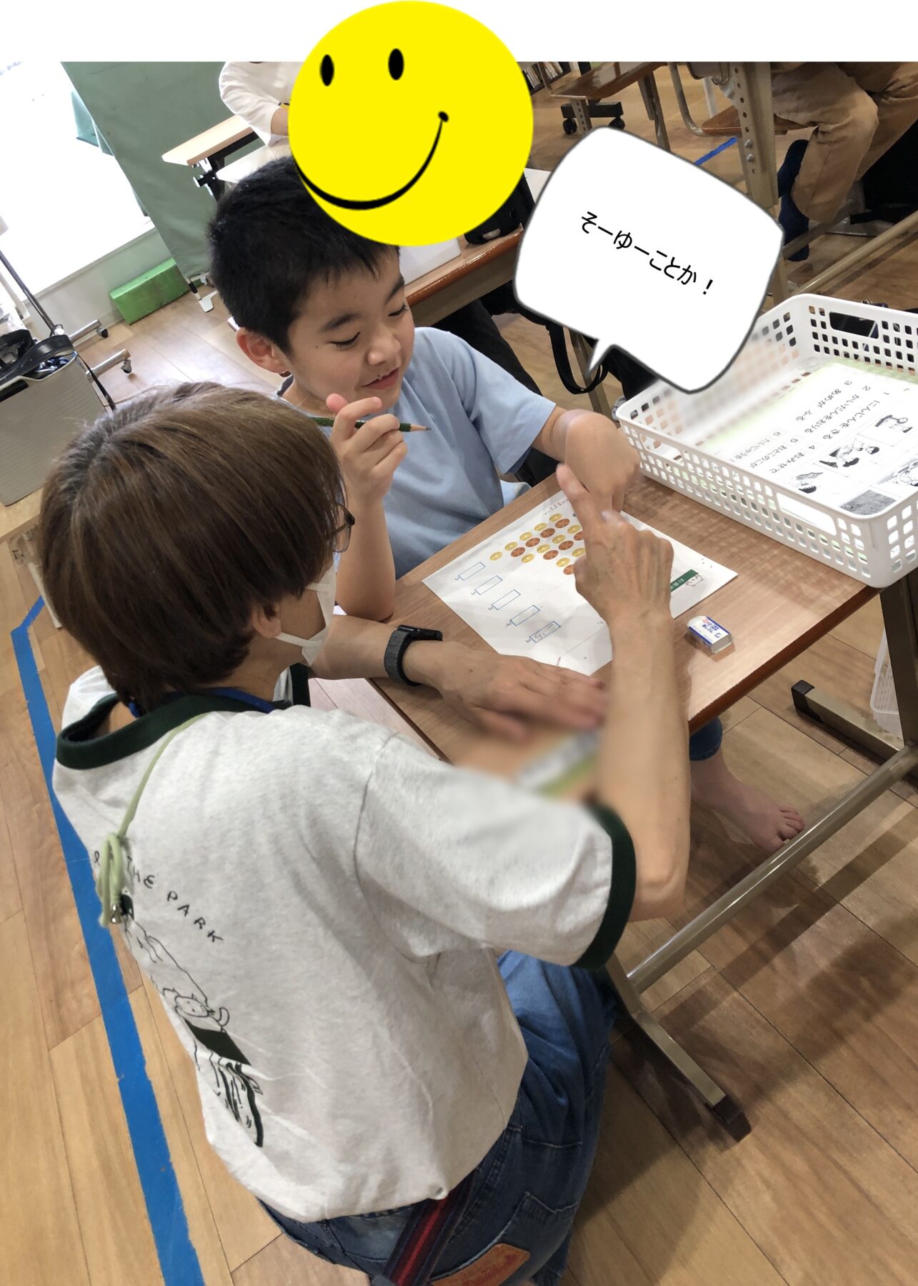 Teacher assists a boy at a desk with a coin-counting worksheet; a large yellow smiley overlay covers part of the scene and a speech bubble reads 'そうゆーことか！'
