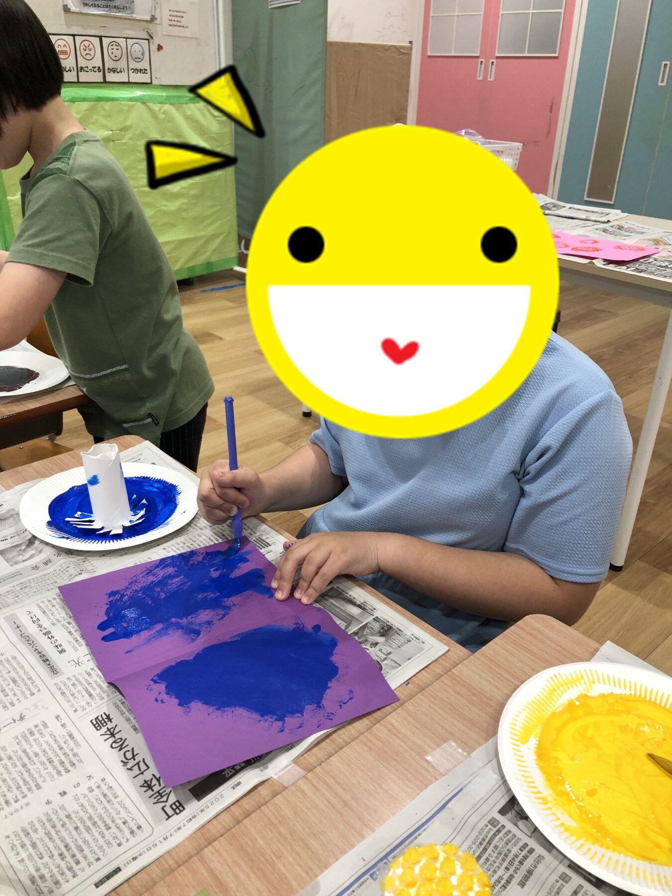 Child in a blue shirt paints blue onto purple paper at a table in a classroom; newspapers spread on the table and colorful room in the background.