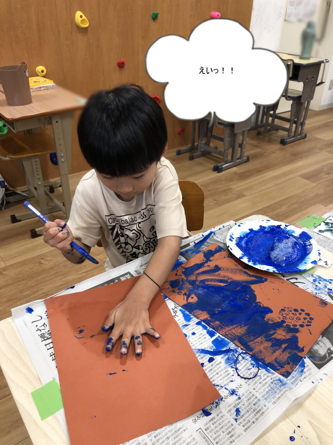 Child in a classroom finger-painting with blue paint on orange paper, hands covered in blue paint; speech bubble reads えいっ！！