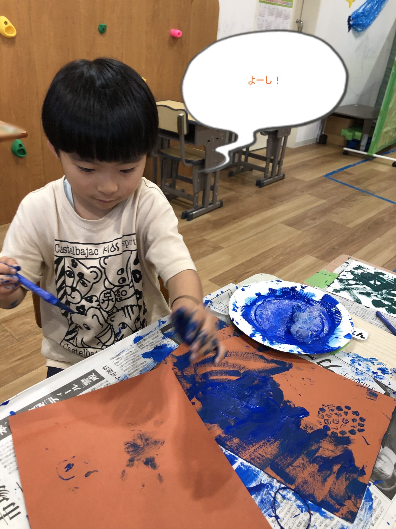 Young child painting at a table, using blue paint on orange paper in a classroom setting, focused on art.