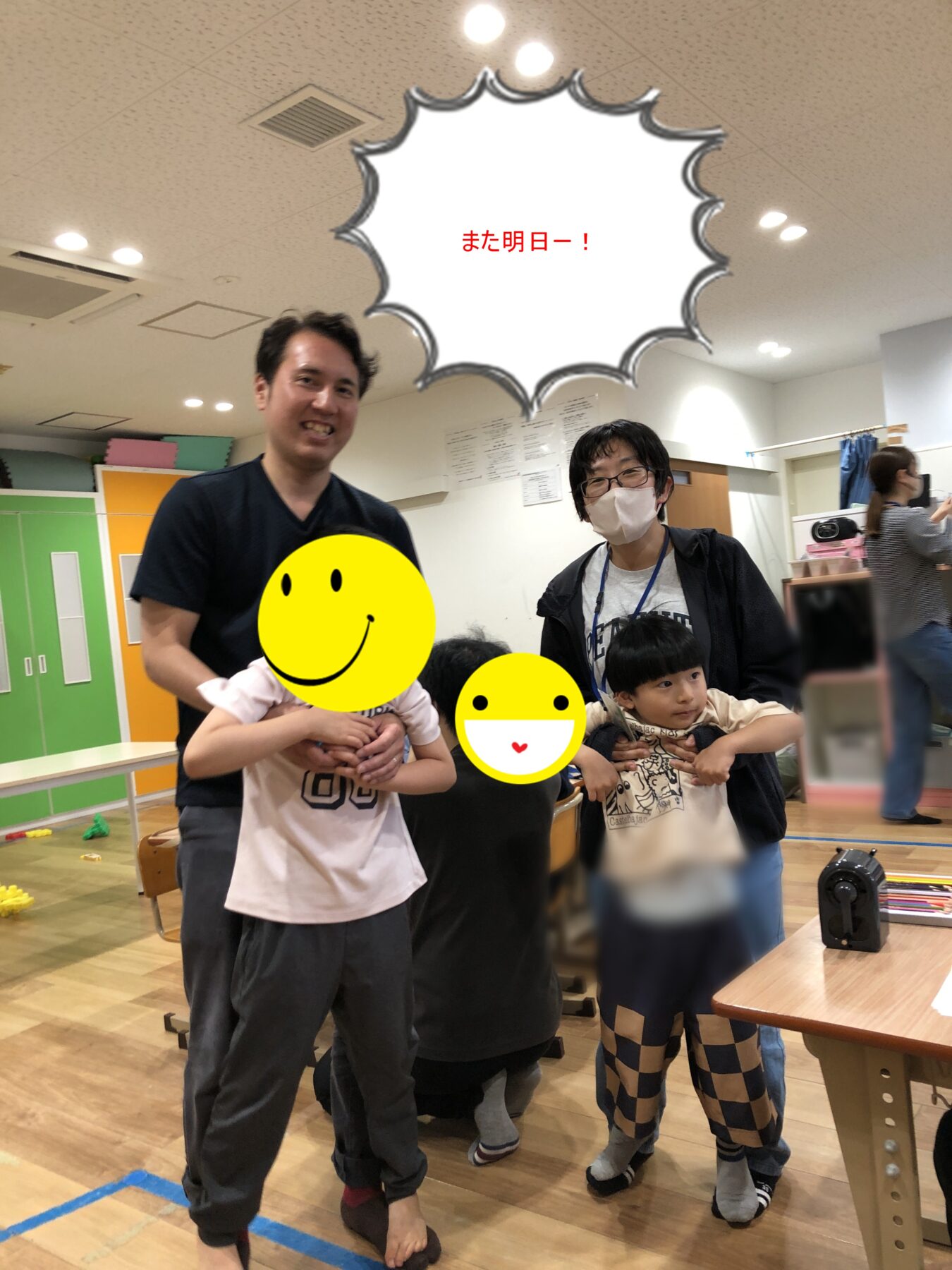 Group of adults and children posing for a photo in a bright classroom; speech bubble shows 'また明日—！' (See you tomorrow).