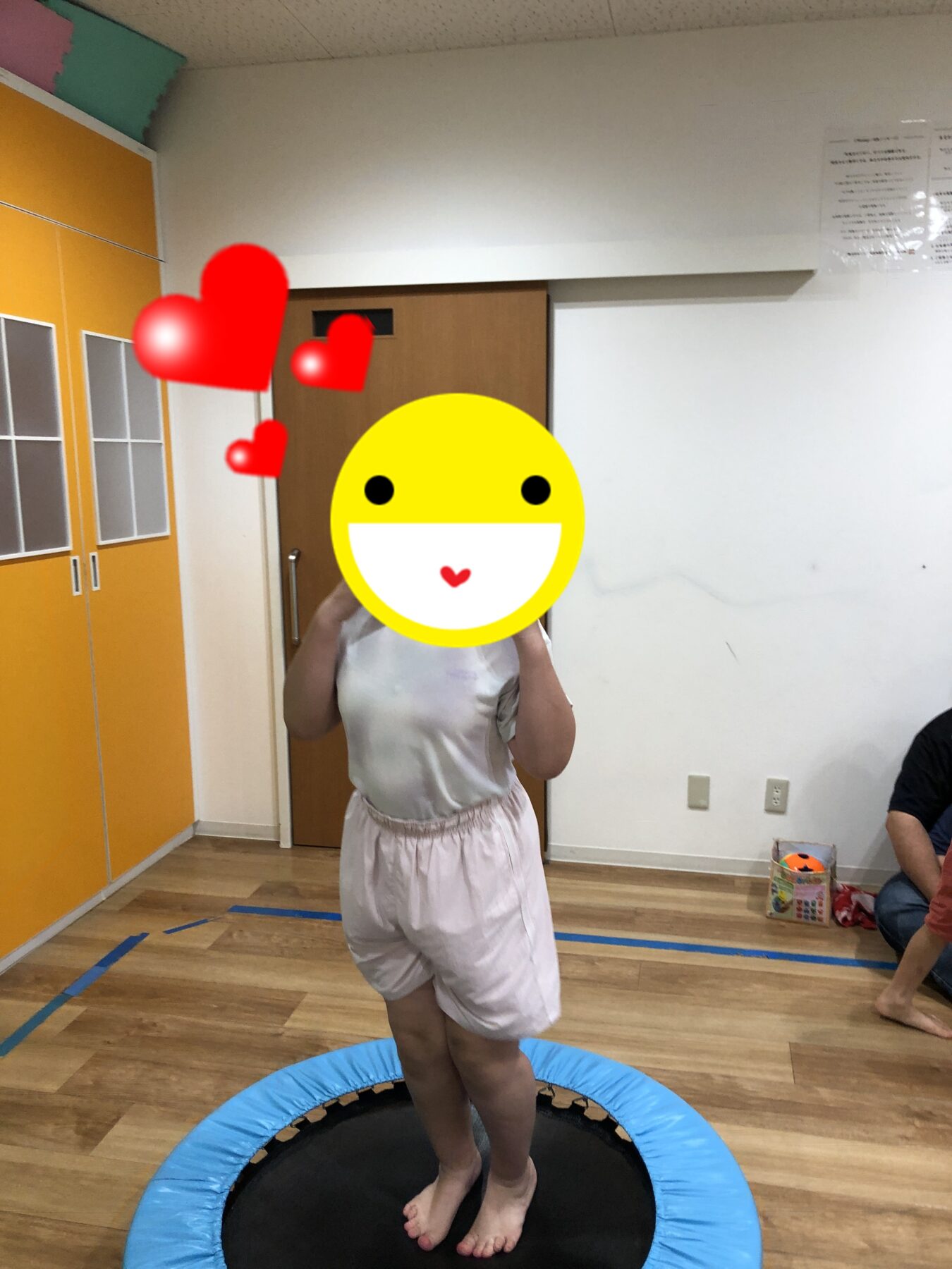 Child stands on a small indoor trampoline, wearing white top and shorts, with a large yellow smiley sticker covering their face, in a room with orange cabinets and wooden floor.