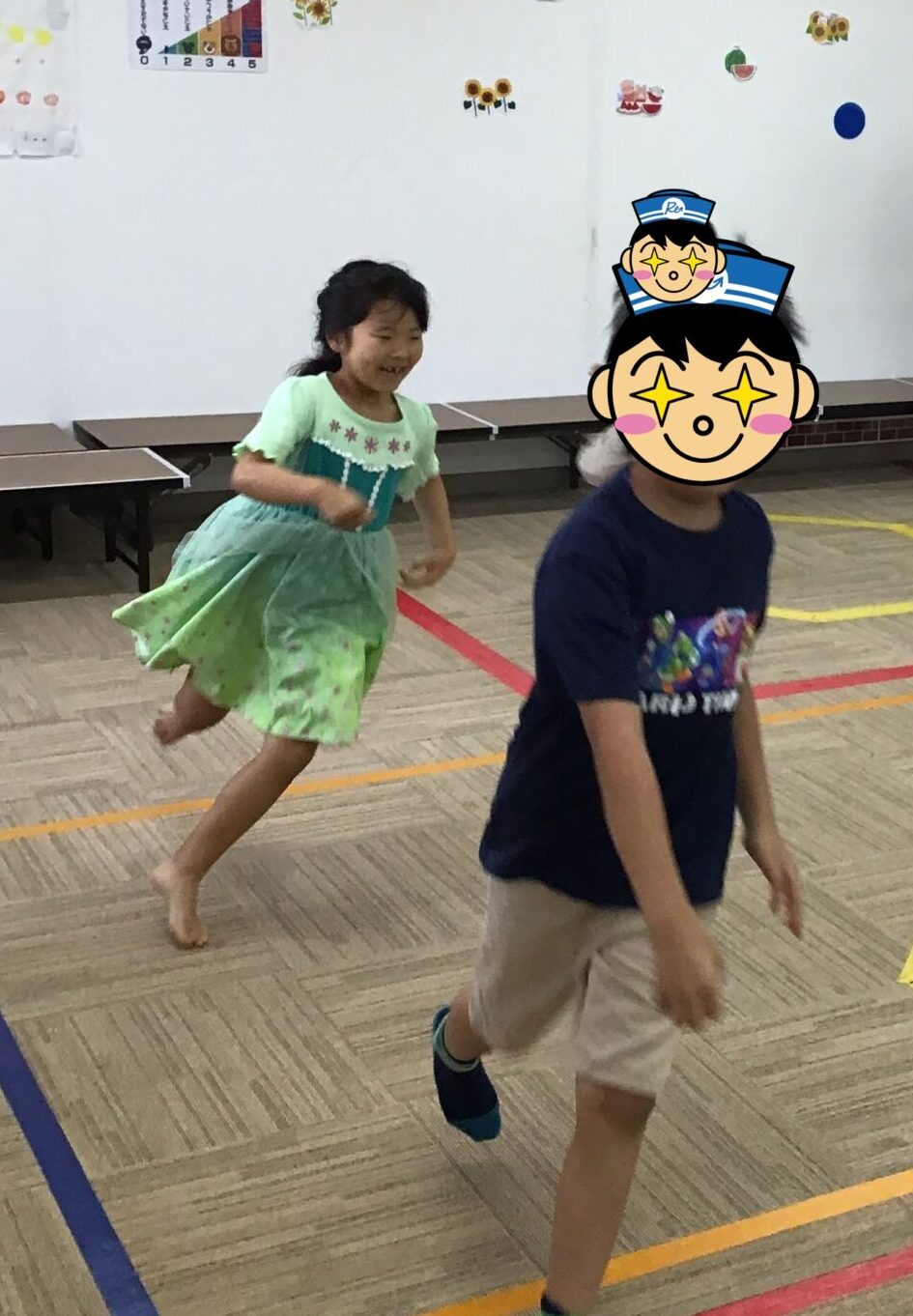 Children running indoors in a gym or large classroom, with a masked adult supervisor standing near the wall and colorful wall decorations.