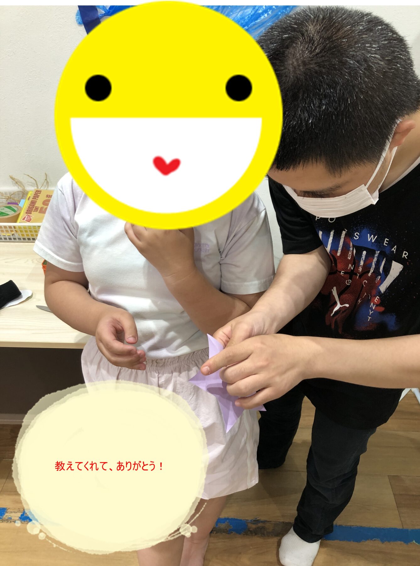 Adult helping a child with a paper craft; the child’s face is covered by a large yellow smiley sticker to protect identity.