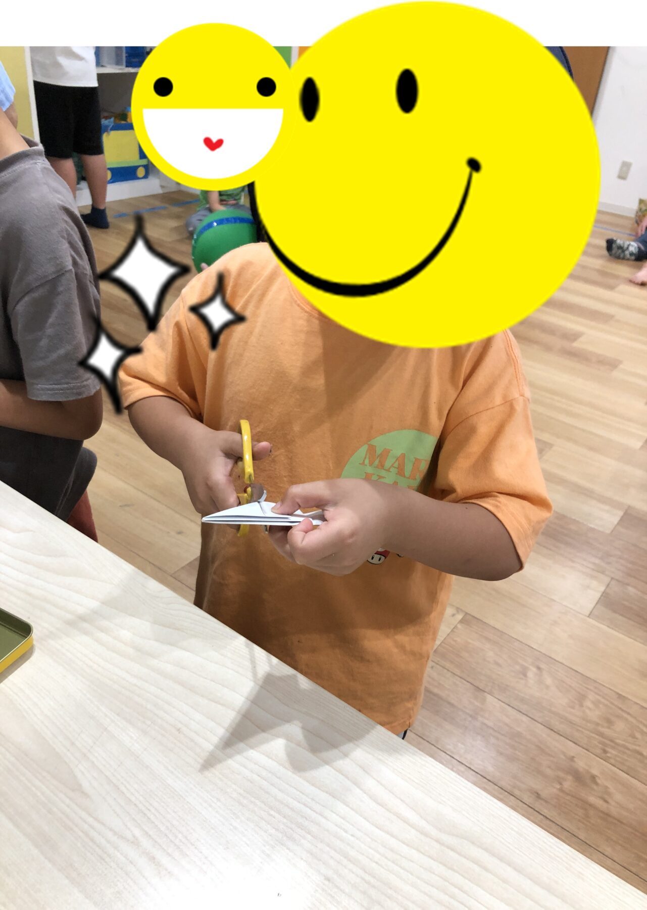 Child cutting paper with yellow scissors at a wooden table in a classroom setting, with cartoon smiley overlays obscuring faces (activity/craft scene).