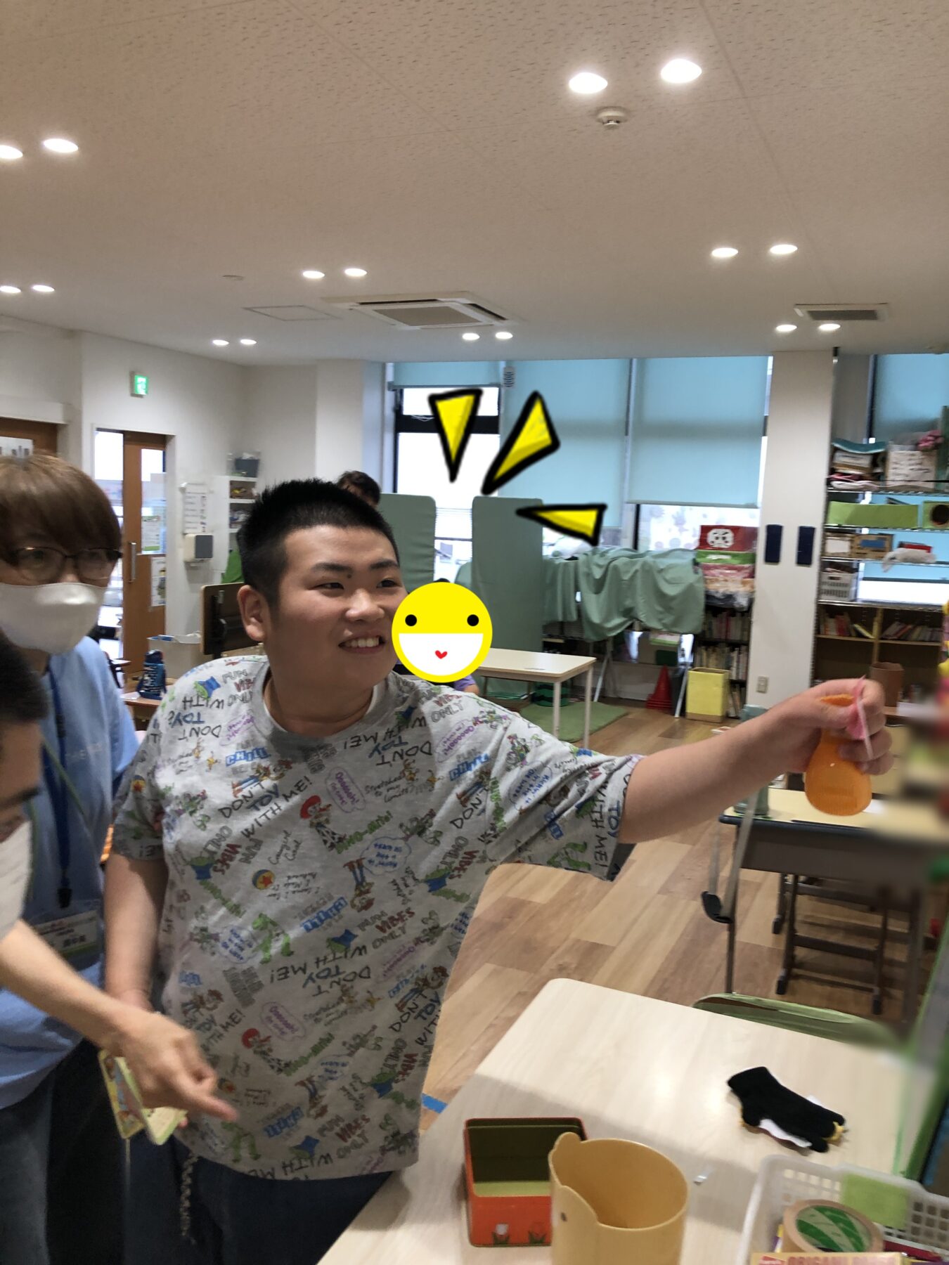 Smiling young person in a colorful graphic shirt stands at a table in a classroom, holding an orange bottle, with another person wearing a mask nearby and craft supplies on the table and shelves in the background.