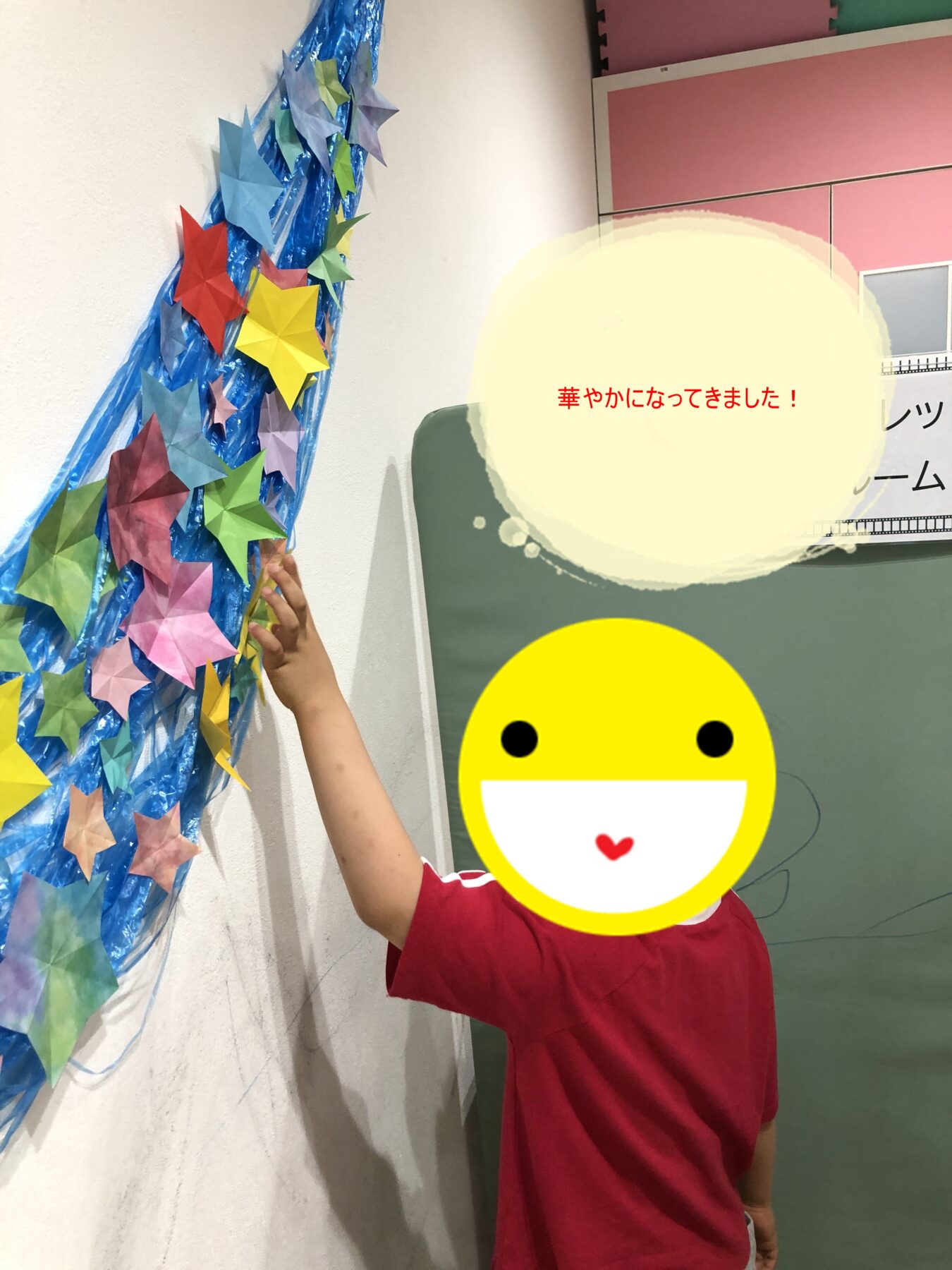 Child in a red shirt places colorful origami stars along a blue streamer wall decoration in a classroom or hallway.