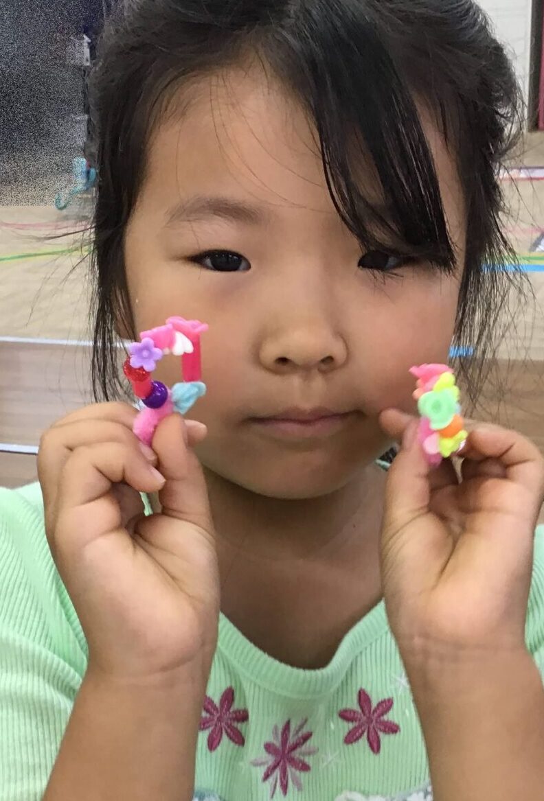 Young girl in a mint-green dress holding two colorful candy-like shapes on sticks up to the camera, smiling gently.