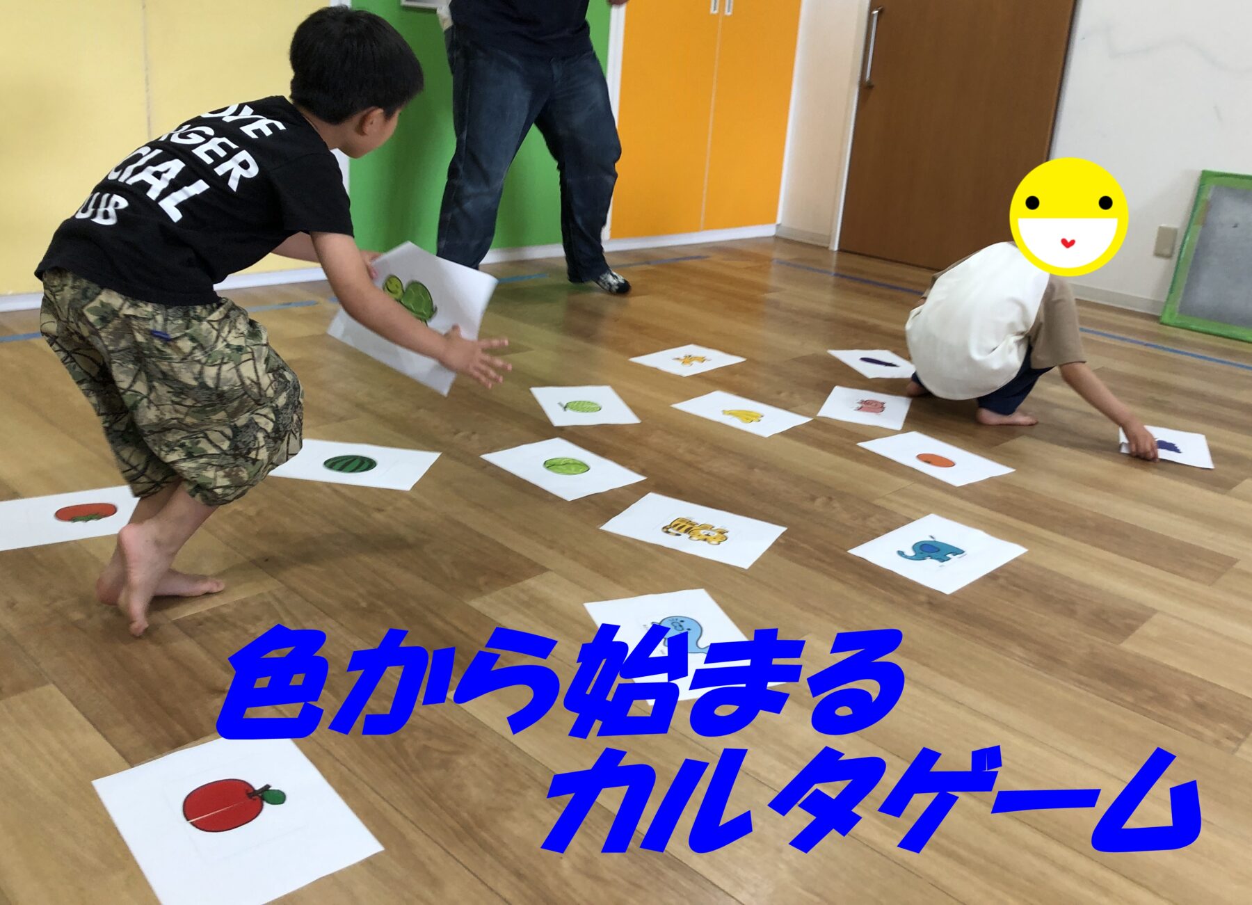 Children playing a floor-based sorting game with printed fruit pictures scattered on white sheets in a brightly colored room. One child is reaching for a card while another sits with a big emoji covering their face.