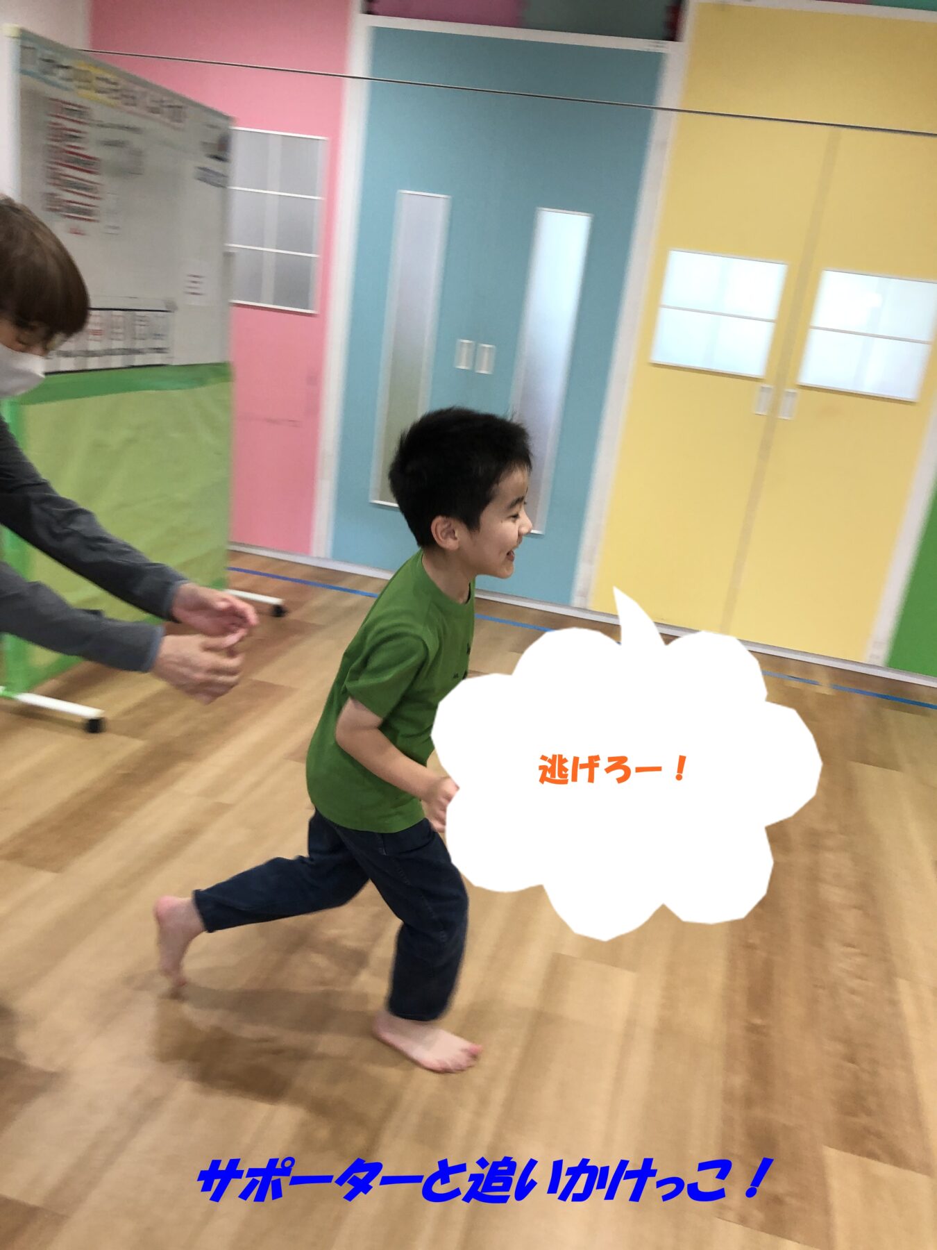 Young boy in a green shirt runs barefoot in a bright indoor playroom as an adult chases him; colorful pink, teal, and yellow walls surround them.