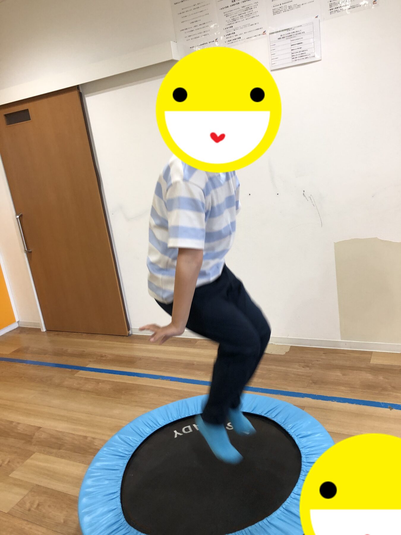 Child mid-jump on a small blue trampoline indoors, wearing a striped shirt; a large yellow smiley mask covers their head.