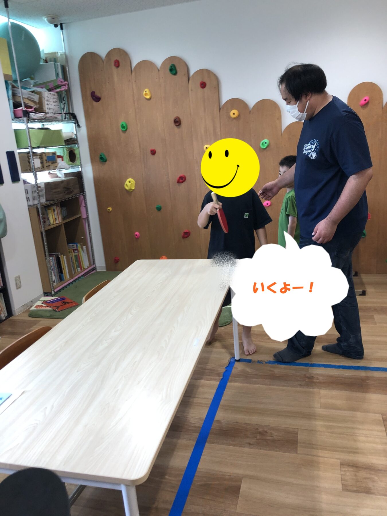 Teacher in a mask helping a child hold a ping-pong paddle beside a wooden climbing wall in a classroom setting.