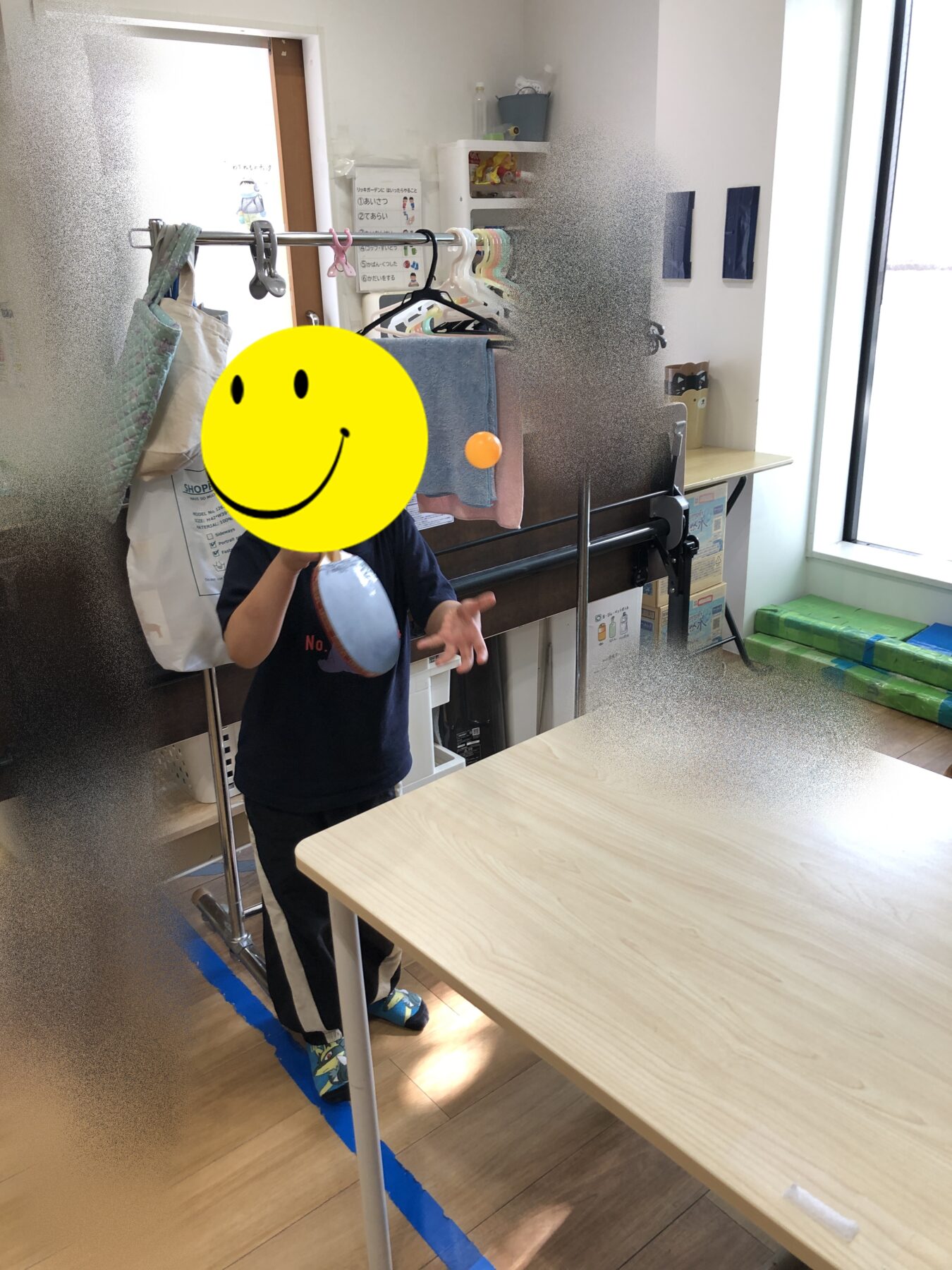 Child standing in a room with a clothing rack and shelves, holding a tambourine in front of their body; face covered by a yellow smiley sticker; blue tape on the floor edge.