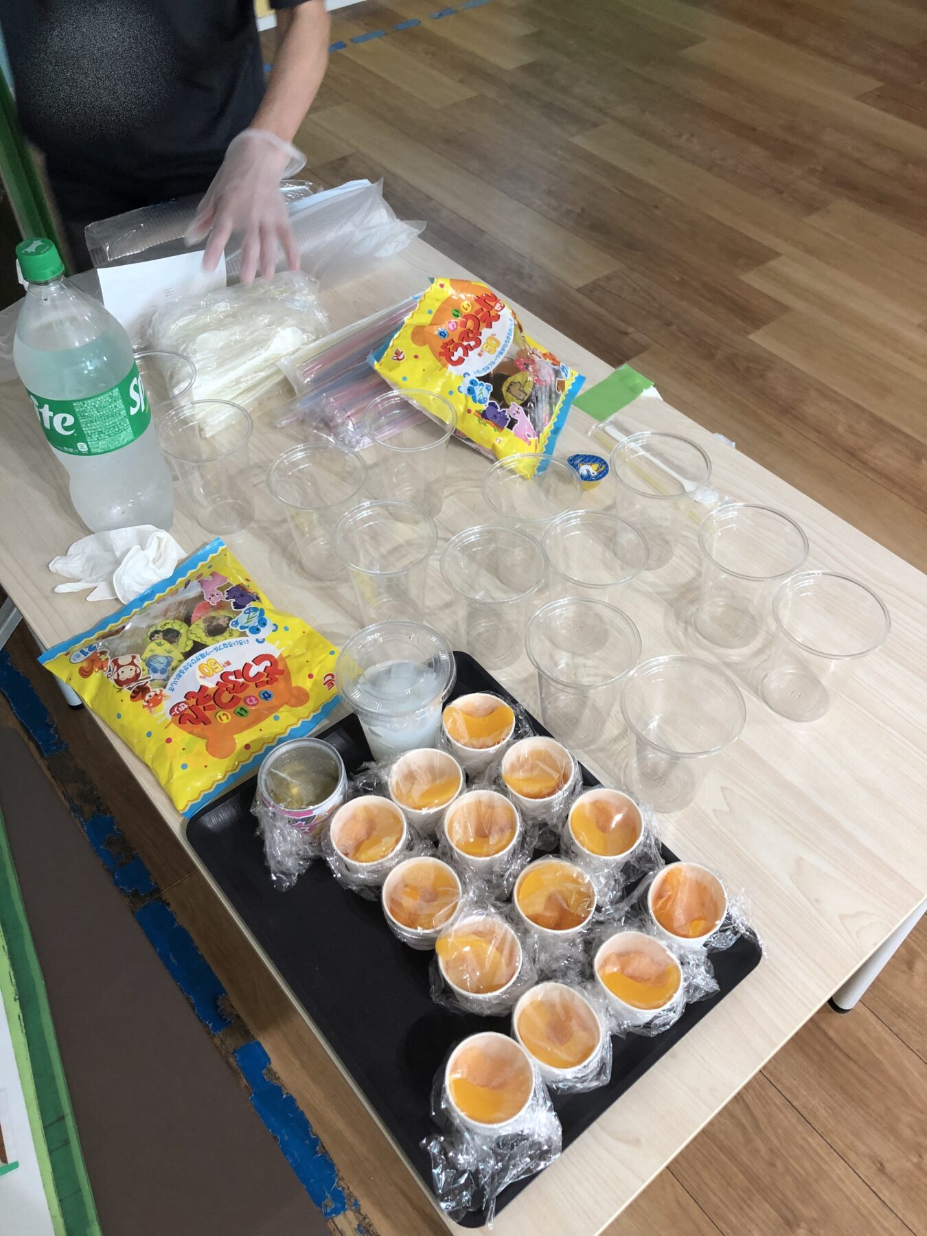 Person wearing a glove arranges clear cups on a wooden table with orange-filled cups and snacks nearby, soda bottle in view.