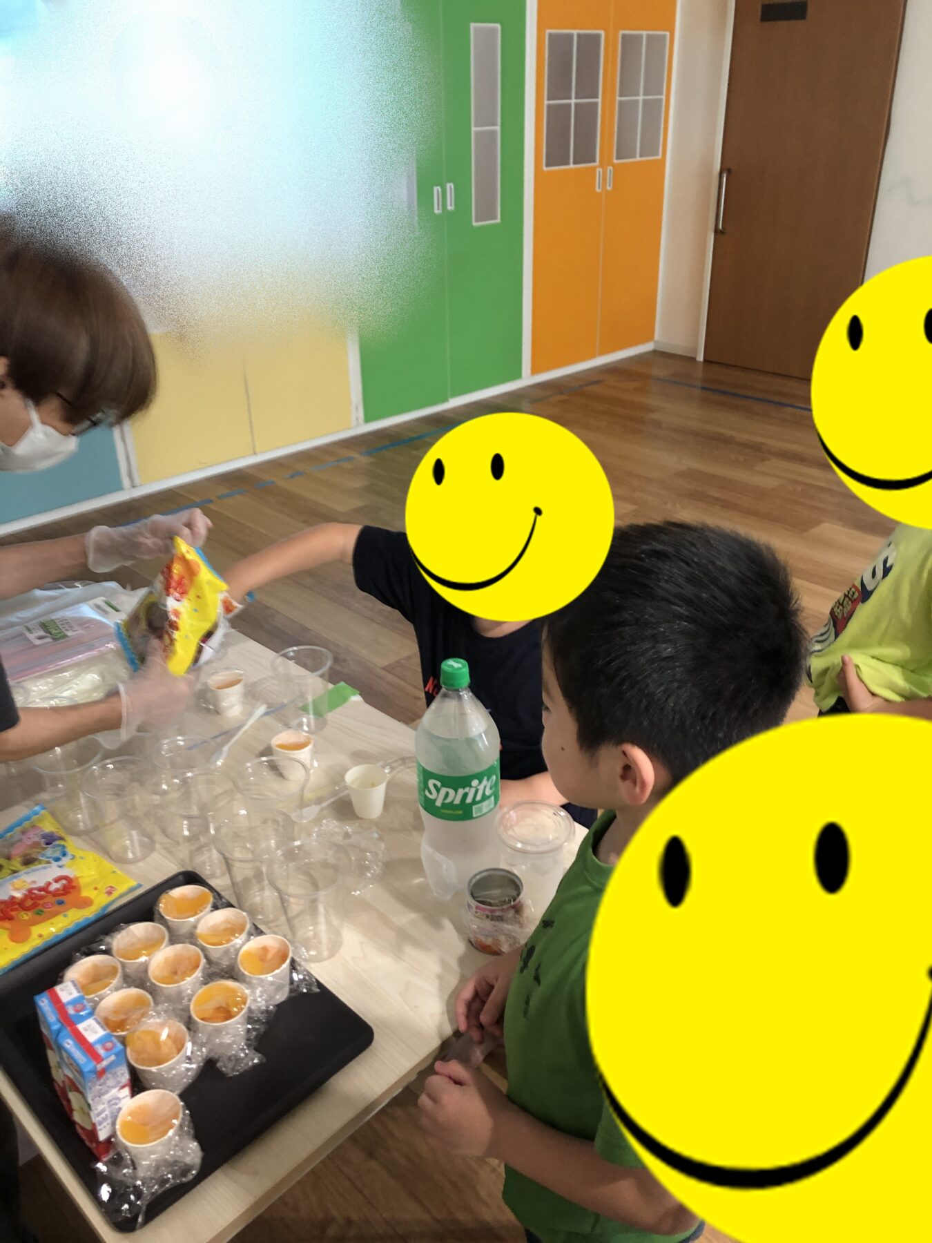 Children sit at a table in a bright classroom while a teacher hands out snacks; cups, a bottle of Sprite, and snack bags are on the table.