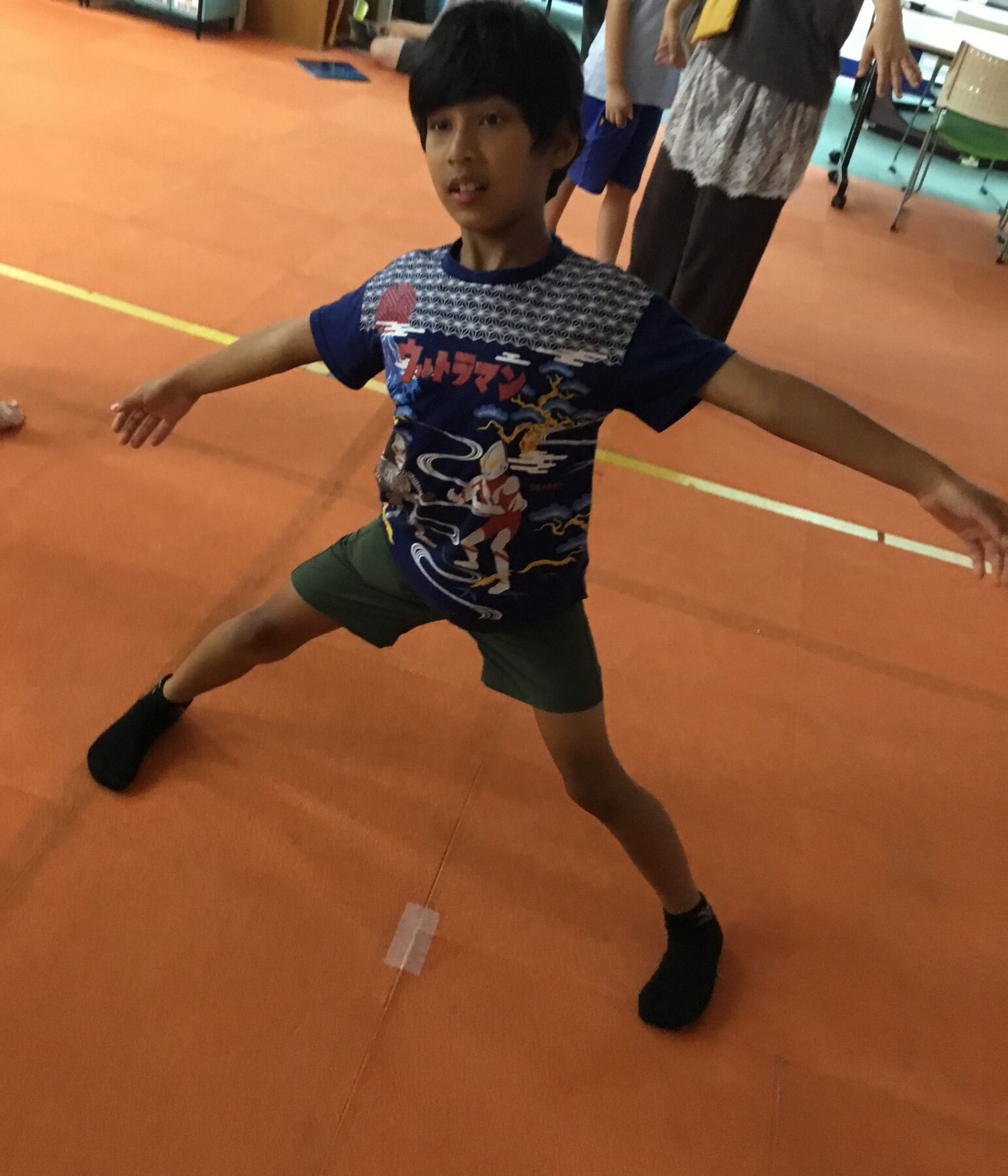Boy in a blue graphic T-shirt and green shorts balancing with arms outstretched in a gymnasium on an orange floor.