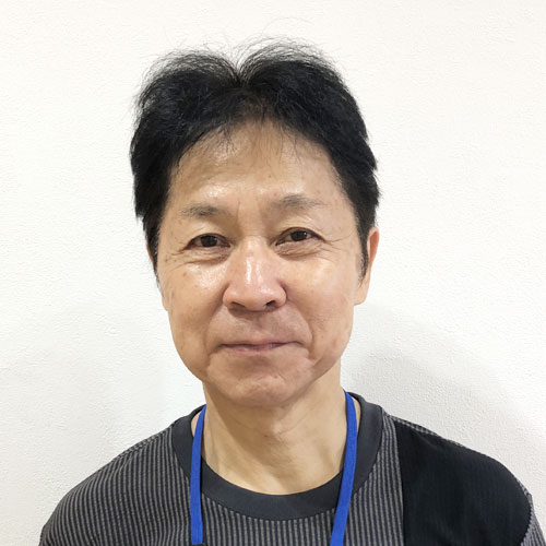Portrait of a middle-aged Asian person with short black hair, wearing a dark striped shirt and a blue lanyard, looking at the camera with a gentle smile.