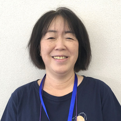 Smiling woman with short black hair wearing a blue lanyard and navy shirt, standing against a light wall.