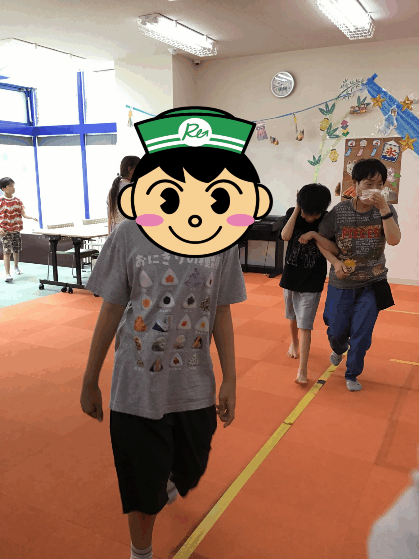 Children walking in a bright activity room with an orange floor; one child in the foreground wears a gray shirt and a large cartoon mask covering their face.