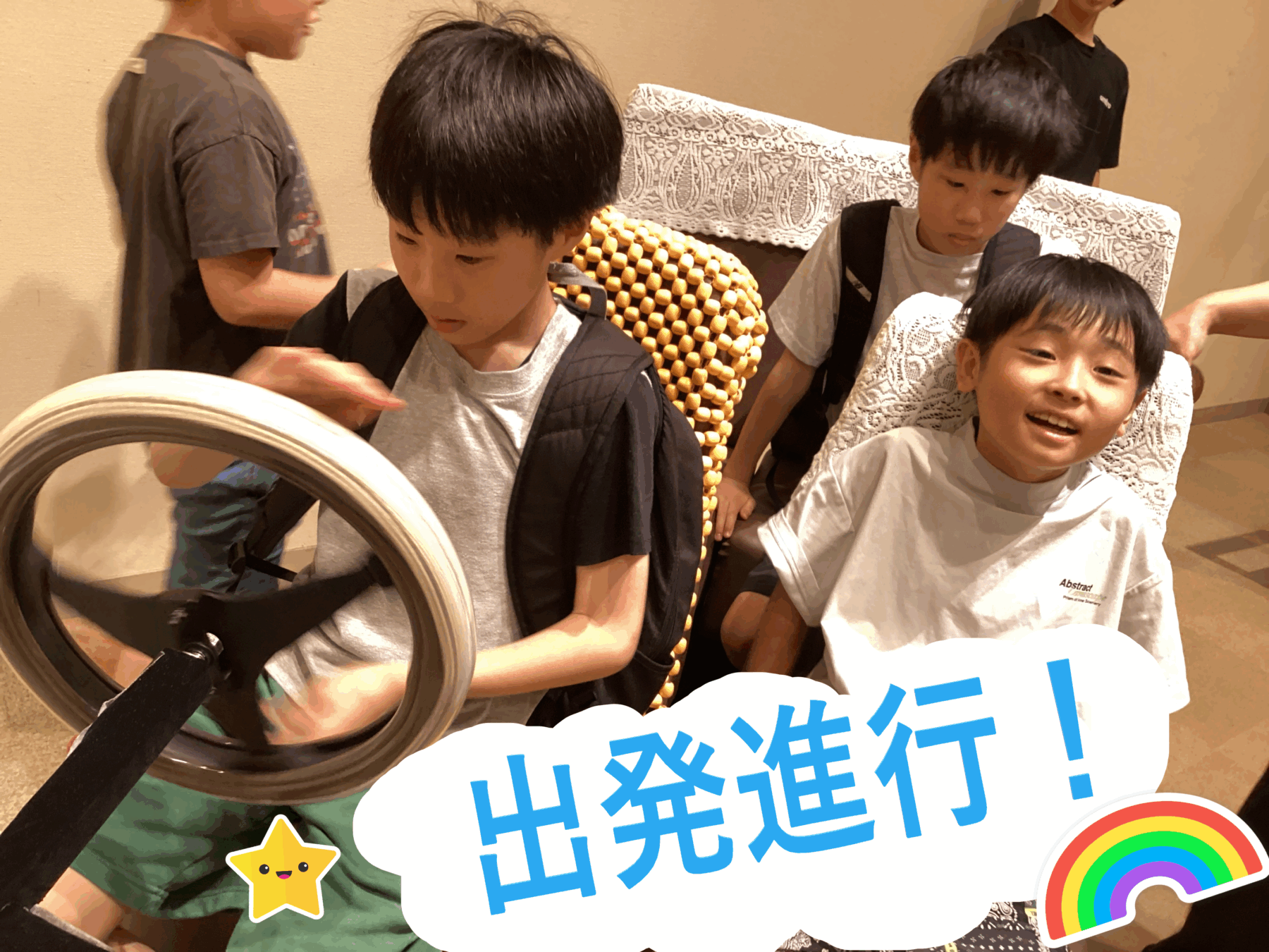 Group of young boys in wheelchairs at an indoor event, smiling and interacting, with colorful stickers (star and rainbow) on a large sign reading '出発進行！' in Japanese.